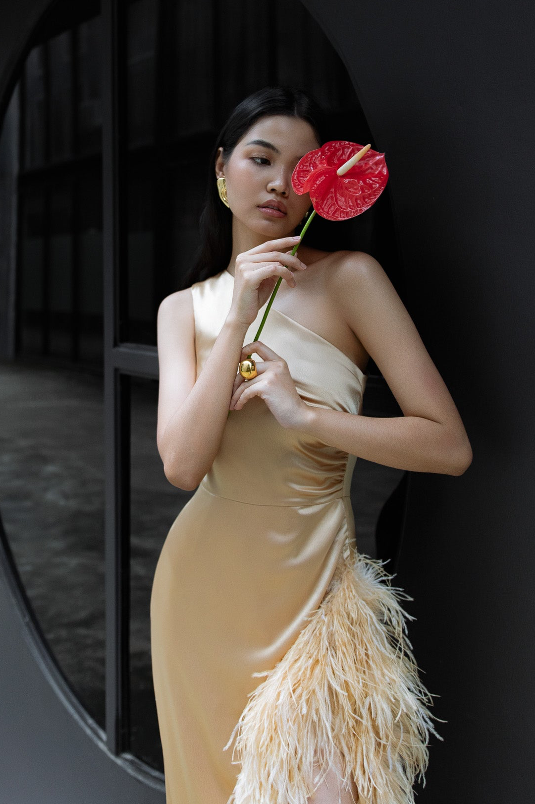 Woman in a feathered beige dress holding a red flower against a dark background. Bangkok Dragontown Photoshoot by Photographer Nadya Shell